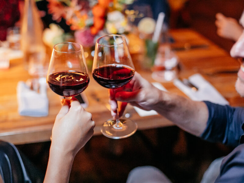 Image has focused forefront and blurred background. It shows the smiling face of a handsome young man with a light beard and mustache clicking glasses with the person sitting to the left of him. The wine glasses contain a red wine. They appear to be at a festive occasion as their are other people seated around the table and a beautiful flower centerpiece in the middle of the table. Image supports our California Chardonnay and California Pinot Noir wine company's blog post about how to talk about wines.