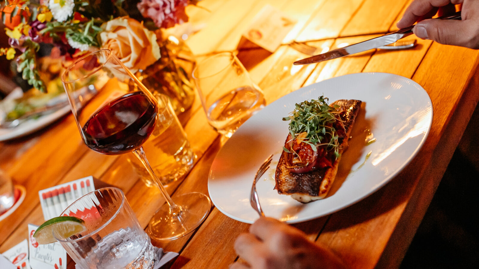 This image shows a beautiful light wood table with beautiful summer flowers in the center, but focuses on the two hands of an individual holding a fork and knife and about to cut into a beautifully garnished piece of roasted fish sitting in front of him. Surrounding the plate of fish are various glasses including one holding pinot noir wine. This image supports our CA pinot noir and chardonnay wine company's blog post about healthy summer recipes and what kind of wine to pair with them.