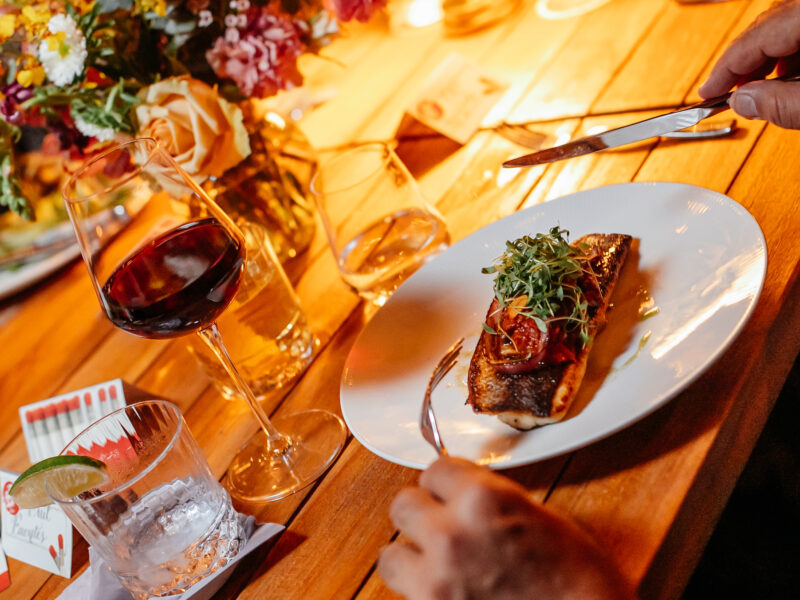 This image shows a beautiful light wood table with beautiful summer flowers in the center, but focuses on the two hands of an individual holding a fork and knife and about to cut into a beautifully garnished piece of roasted fish sitting in front of him. Surrounding the plate of fish are various glasses including one holding pinot noir wine. This image supports our CA pinot noir and chardonnay wine company's blog post about healthy summer recipes and what kind of wine to pair with them.
