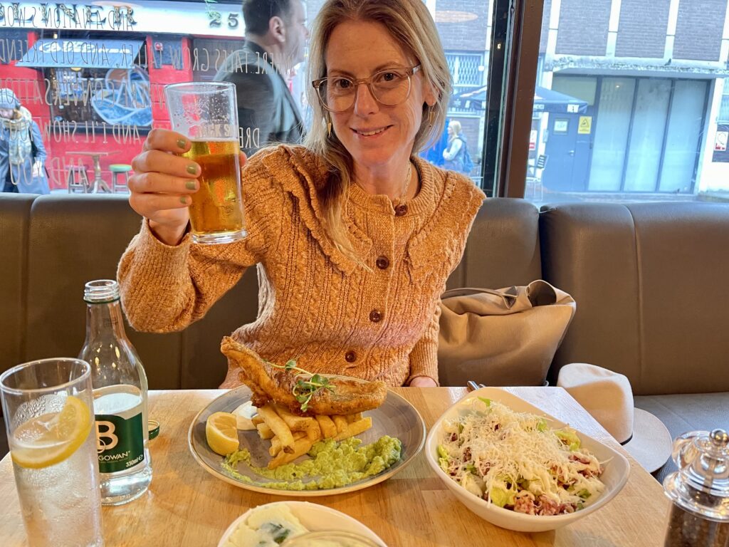 A woman smiles and holds up a glass of beer. A plate of fish and chips sits in front of her.