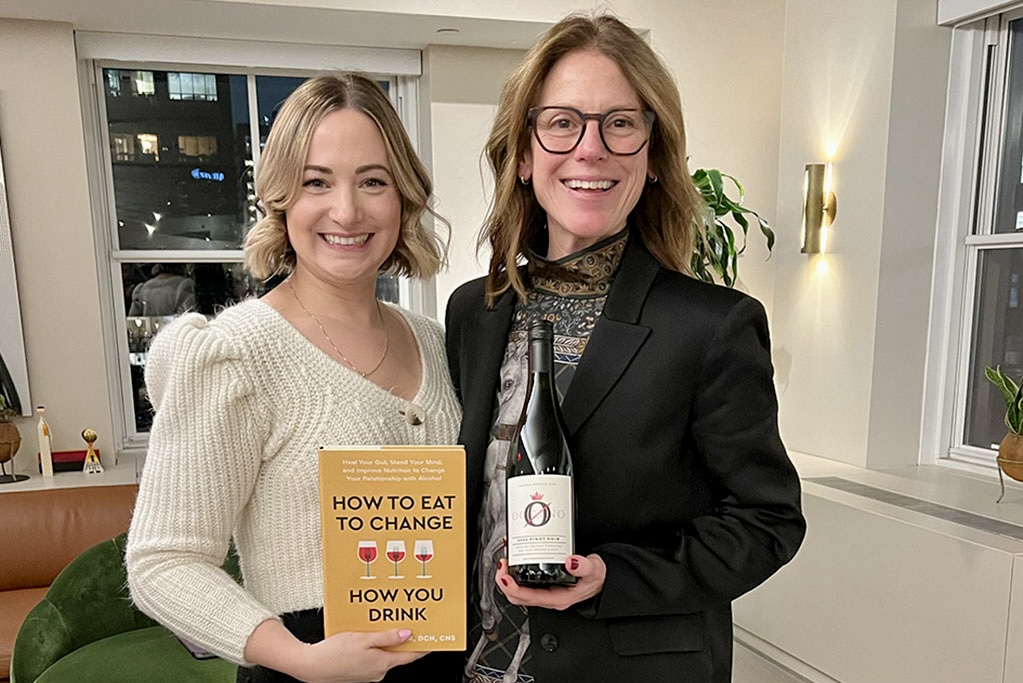 Two smiling women, one holding the book How to Eat to Change How You Drink and one holding a bottle of alcohol free wine.