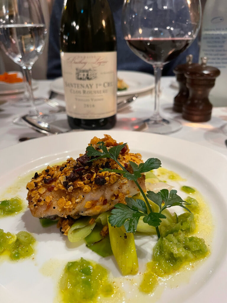 A meal served on a white plate sits on a white table in front of glass and bottle of red wine.