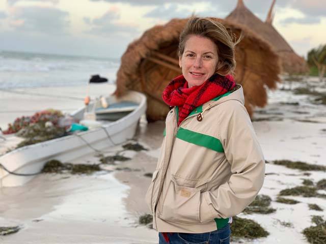 Rachel Martin, owner of Oceano Wines, standing on a beach on Isla Holbox, next to a boat and a small hut with a thatched roof.