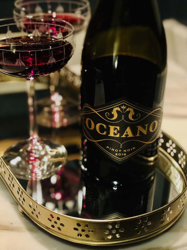 A bottle of Oceano Wines' 2018 Pinot Noir resting on a mirrored tray next to a glass of red wine.