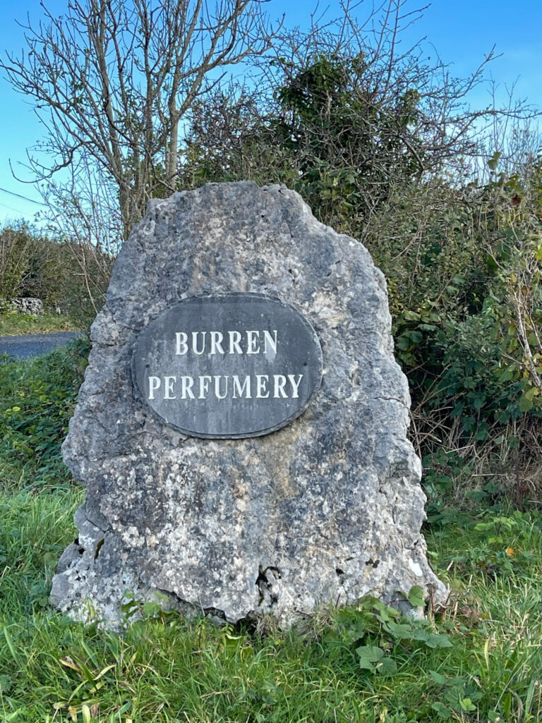 A Burren Perfumery sign on a rock stands in front of bushes and trees.