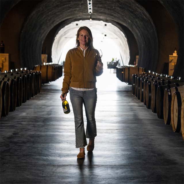 A woman carrying a wine bottle in one hand and glass in the other walks down a path lined with wine barrels.