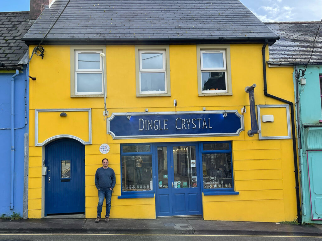 A man in a blue sweater and jeans stands in front of a yellow building with a blue door and a sign that says "Dingle Crystal."