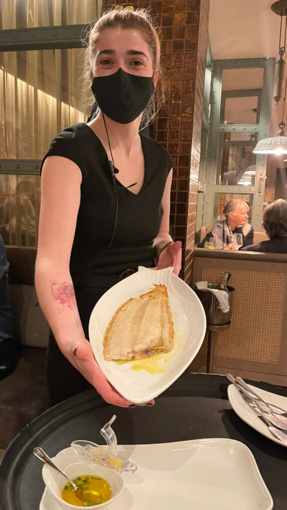 A woman wearing a black dress and black face mask tilts a plate of fish toward the camera.