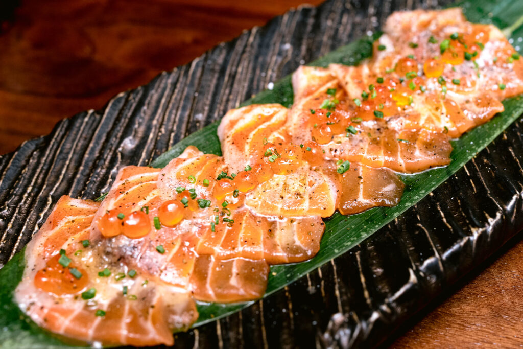 Fish topped with fish eggs and chives over a green leaf on a black plate.