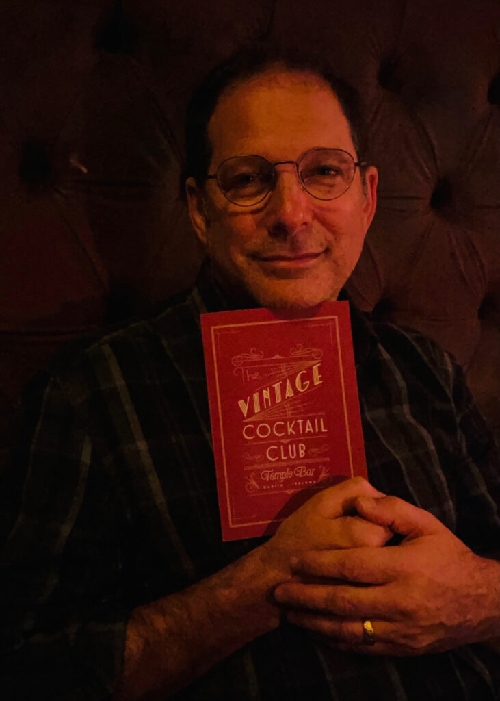 A man holds a red paper that reads "The Vintage Cocktail Club" and smiles.