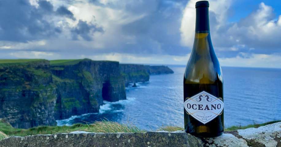 A bottle of Oceano wine sits on a rock wall overlooking water and cliffs.