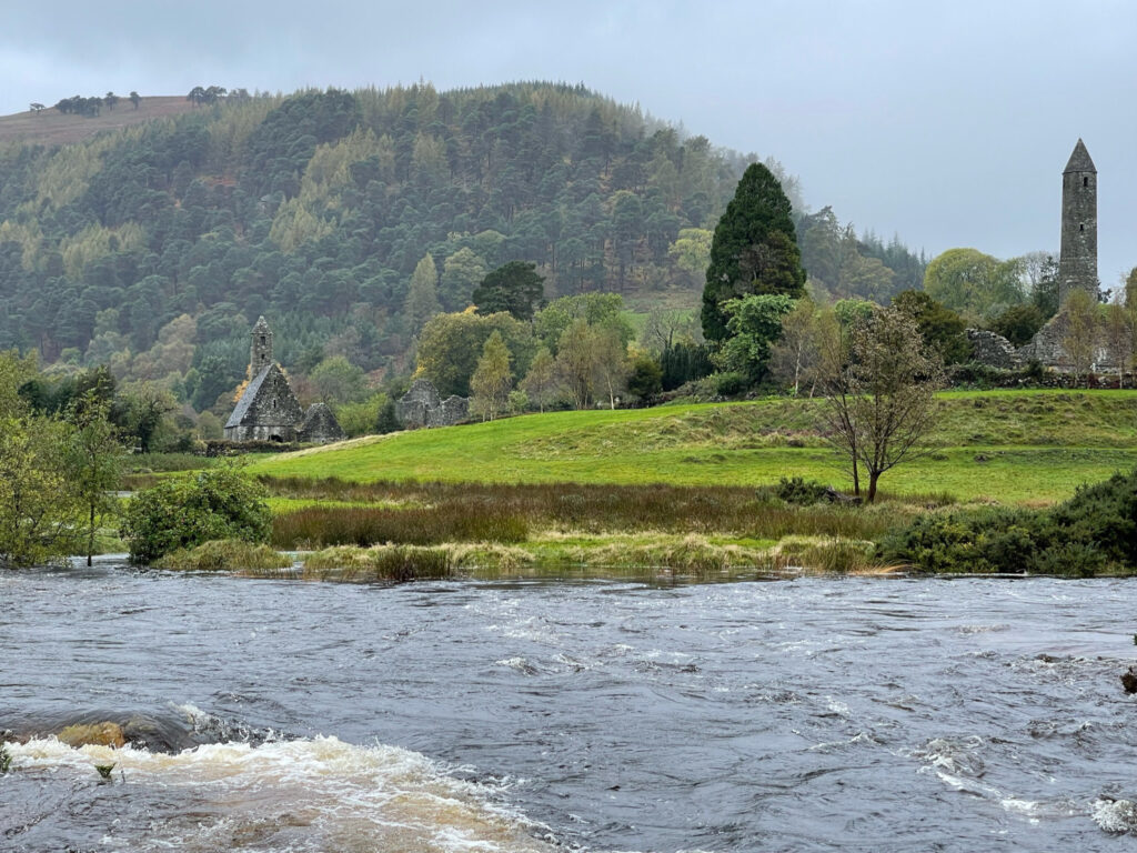 Running water in front of a patch of green grass, a stone building, and a hill with trees on it.