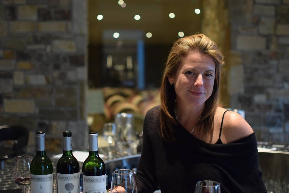 A smiling woman sits next to three wine bottles and several wine glasses.