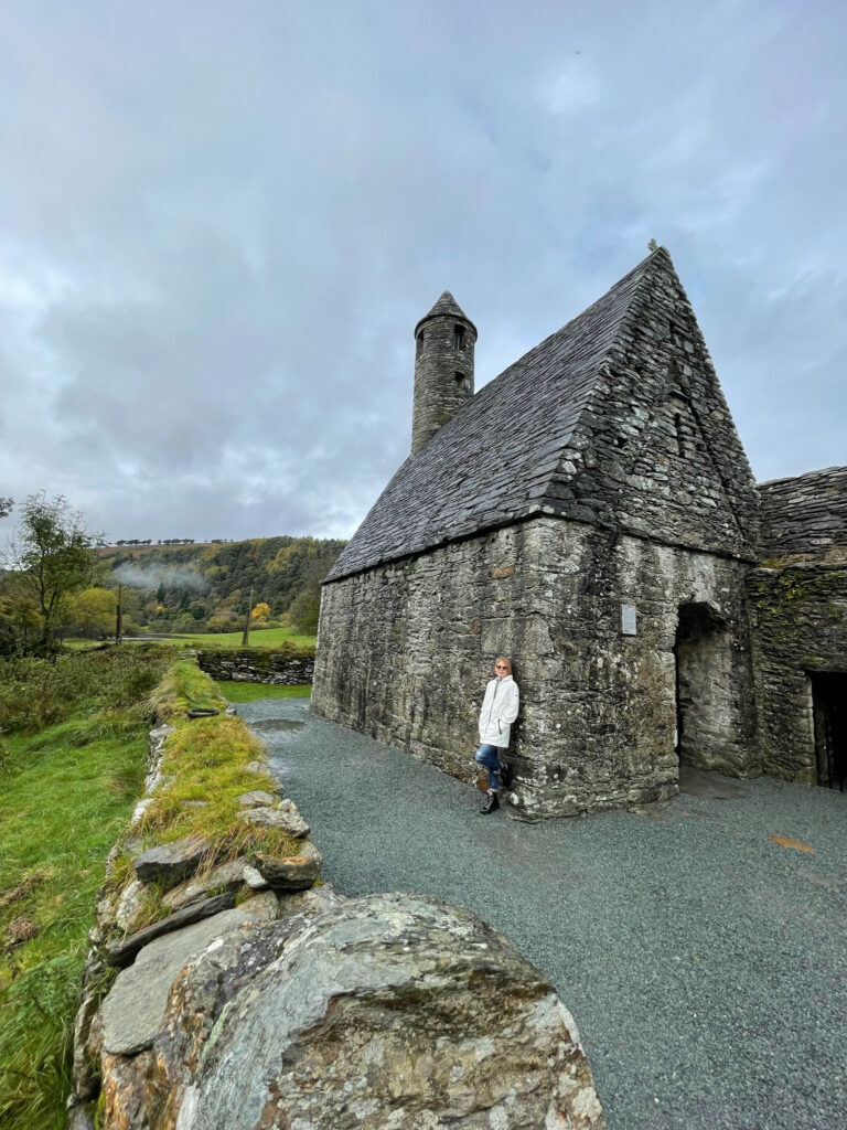 A woman in a white coat leans against a stone building.