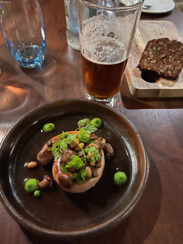 A sausage sits on a brown plate next to a glass of beer.