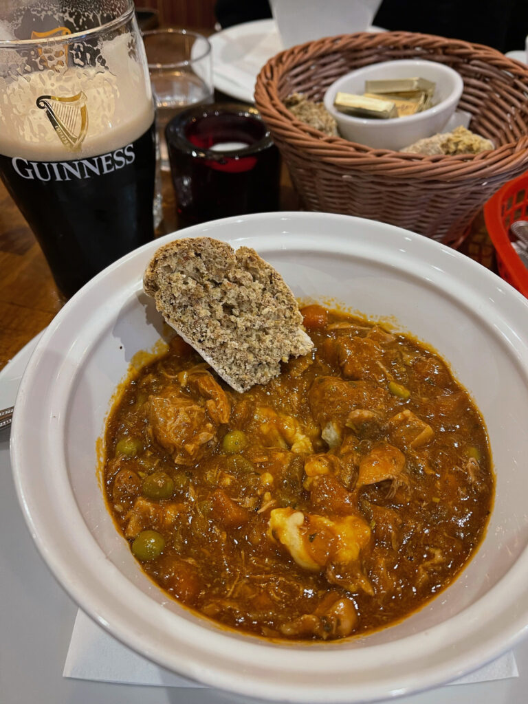 A piece of bread sits in a bowl of stew next to a bread basket and a glass of Guinness beer.