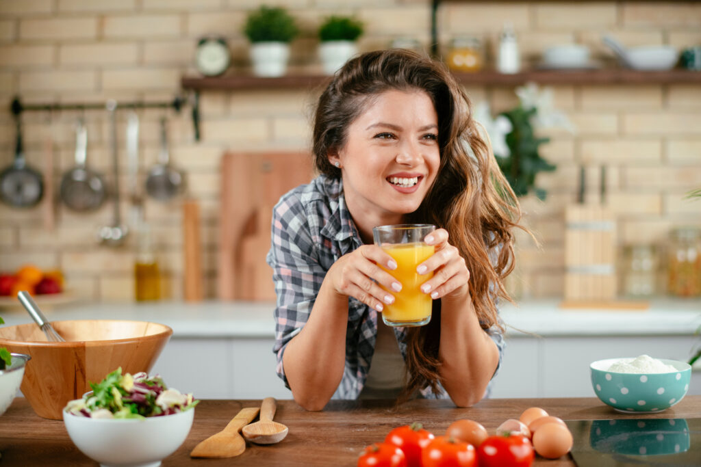 A woman smiles and leans against a kitchen counter while holding a glass of juice. There are bowls on the counter.