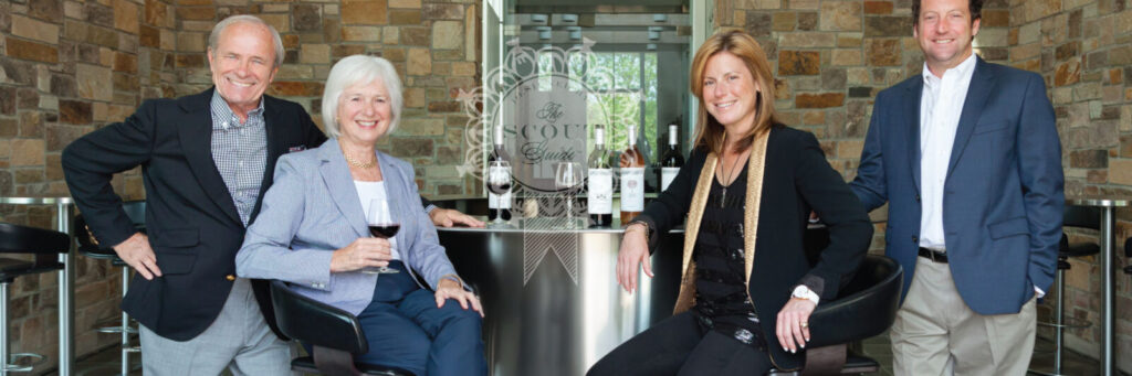 Two men stand next to two women sitting in bar stools next to a bar with wine glasses and bottles.