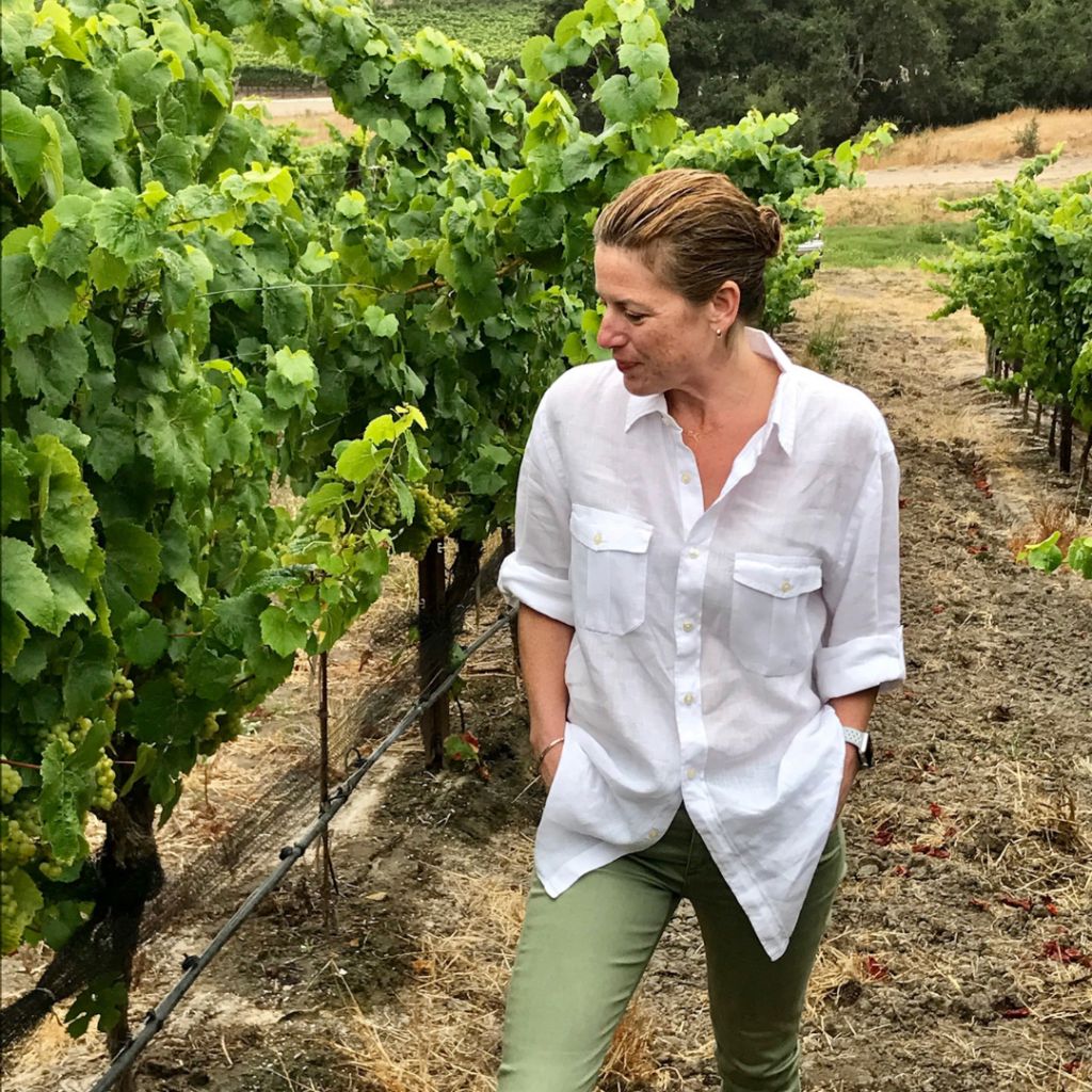 A woman in a white shirt and green pants looks at grape vines.