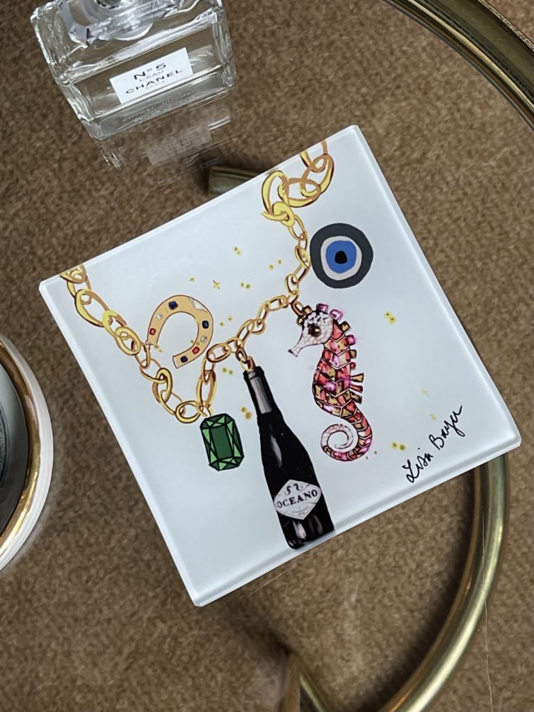 A small white tile with a seahorse, a wine bottle, a horseshoe, and an emerald painted on it.