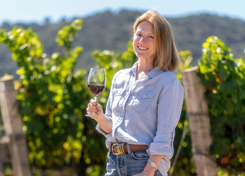 A woman holding a glass of non-alcoholic red wine in front of her vineyard on the San Luis Obispo coast.