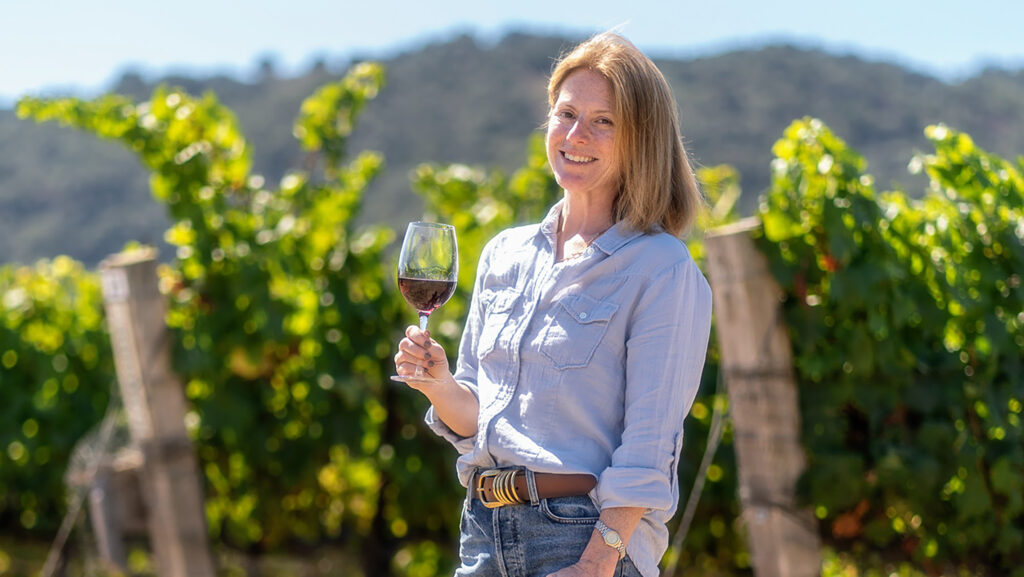 A woman holding a glass of non-alcoholic red wine in front of her vineyard on the San Luis Obispo coast.