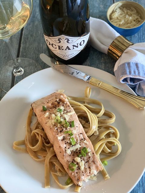 A plate of pasta and a protein next to a bottle of Oceano's Chardonnay wine.