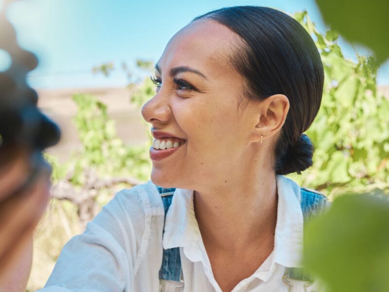 A woman smiles and looks to her side as she picks grapes off a vine on a sunny day.