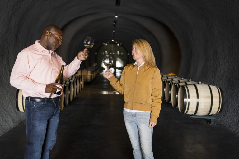 A man and woman hold glasses of wine up to one another in a wine cellar.