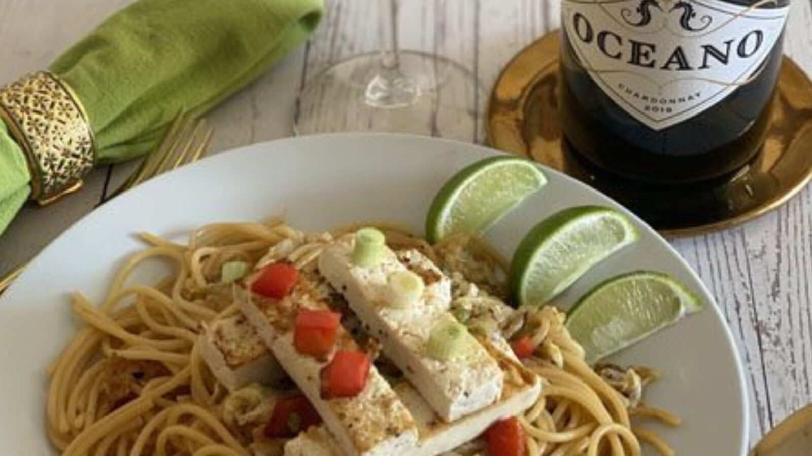 A plate with a tofu and noodle dish and three limes next to a wine bottle and green fabric napkin.