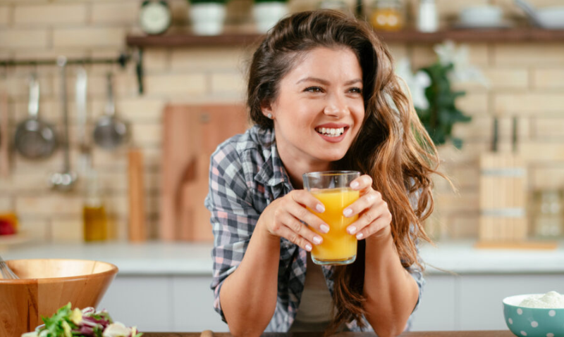 A woman smiles and leans against a kitchen counter while holding a glass of juice. There are bowls on the counter.