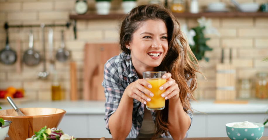 A woman smiles and leans against a kitchen counter while holding a glass of juice. There are bowls on the counter.