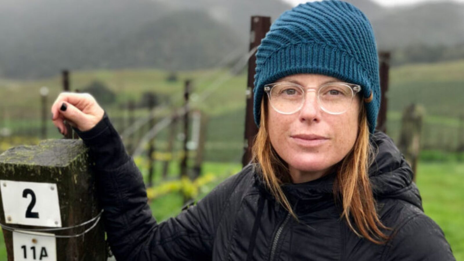 Rachel Martin, founder of Oceano Wines, standing in front of a large vineyard and wearing a blue beanie.