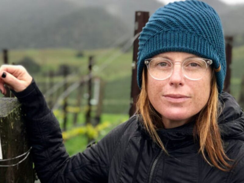 Rachel Martin, founder of Oceano Wines, standing in front of a large vineyard and wearing a blue beanie.