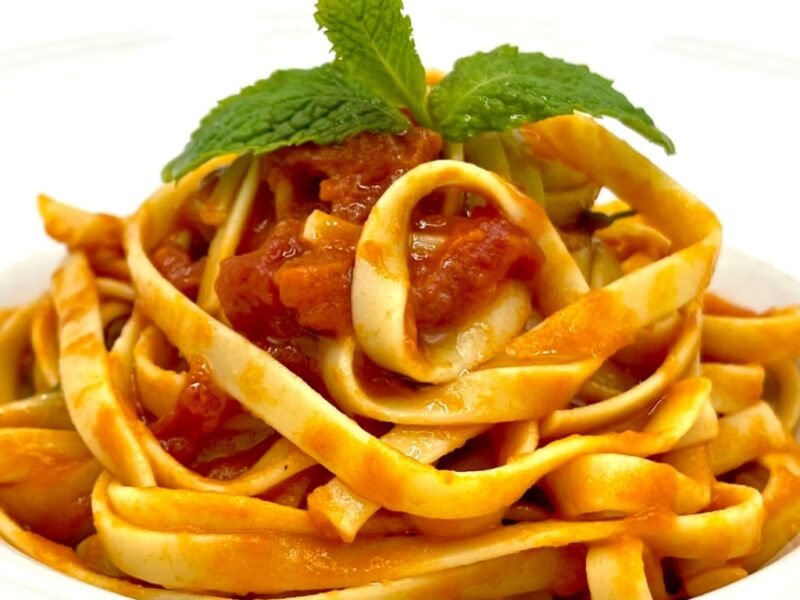 A helios kordonia pasta with fresh tomato sauce on a white plate, topped with a sprig of basil.