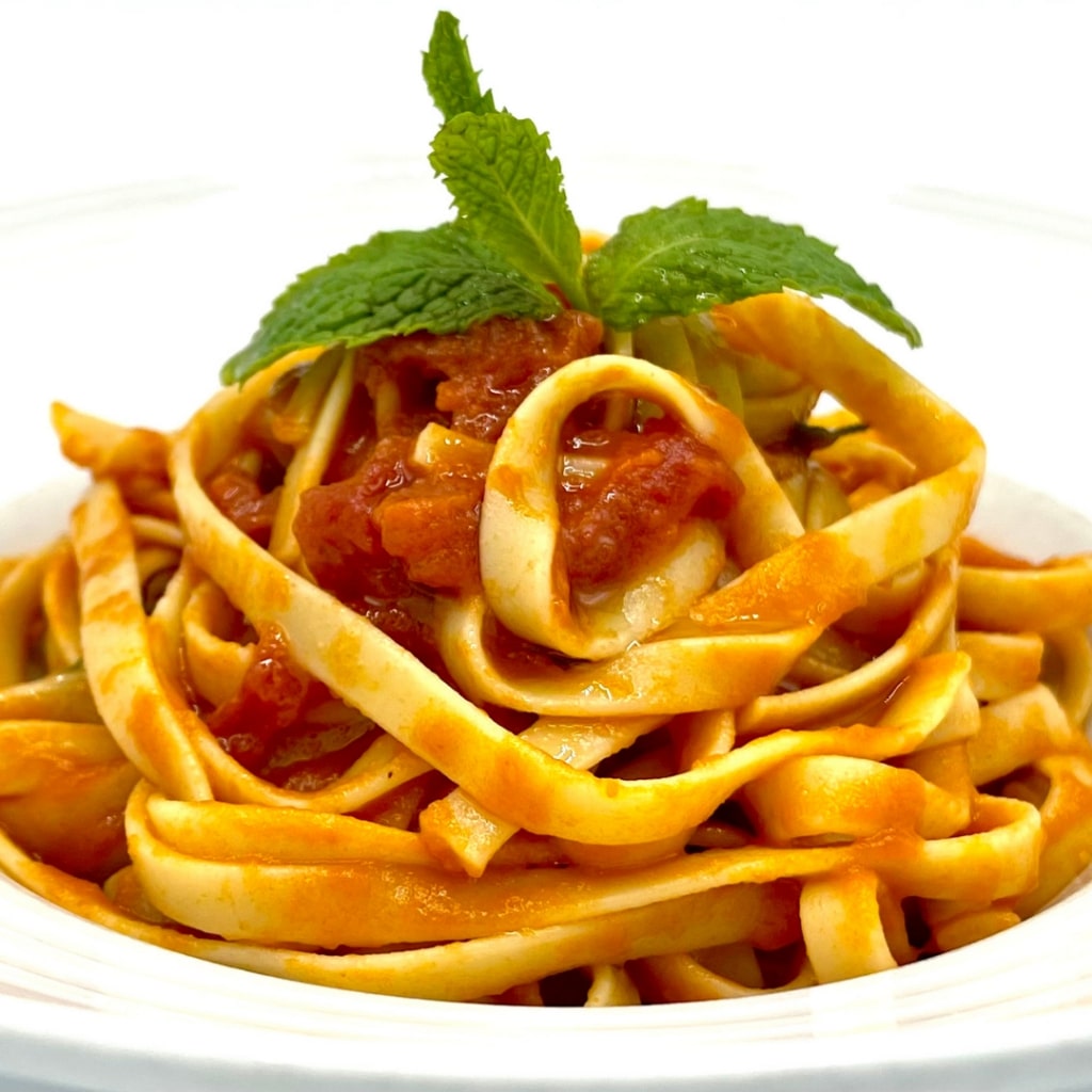 A helios kordonia pasta with fresh tomato sauce on a white plate, topped with a sprig of basil.