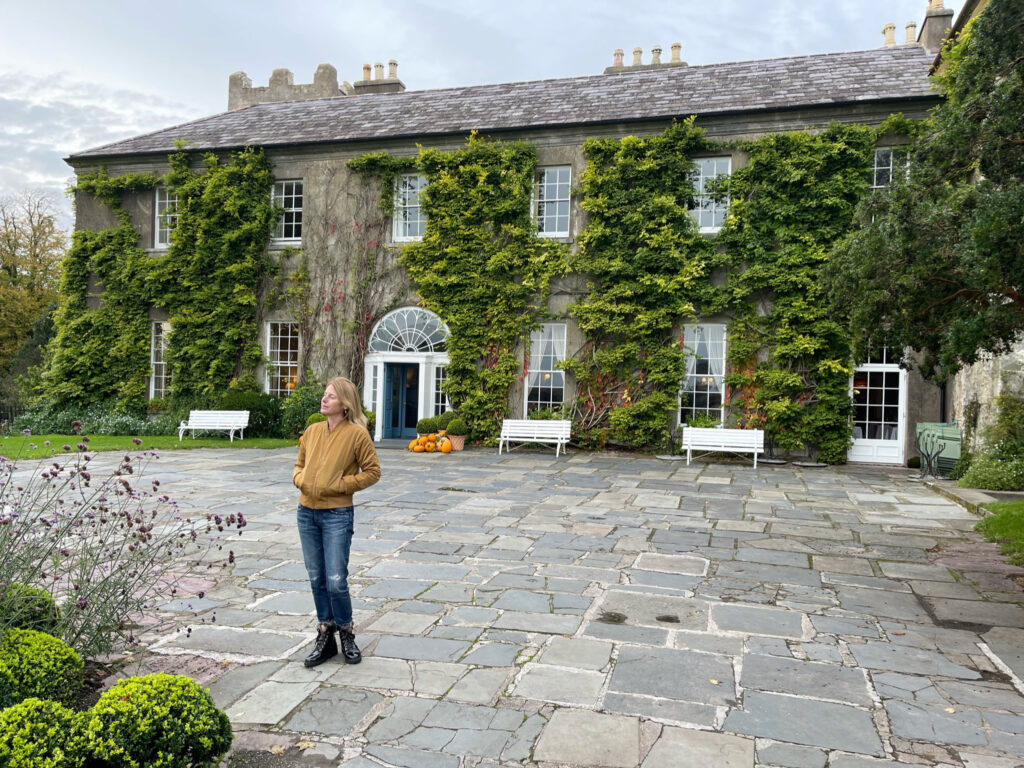 A woman in a yellow coat stands in front of a stone building with ivy growing on it.