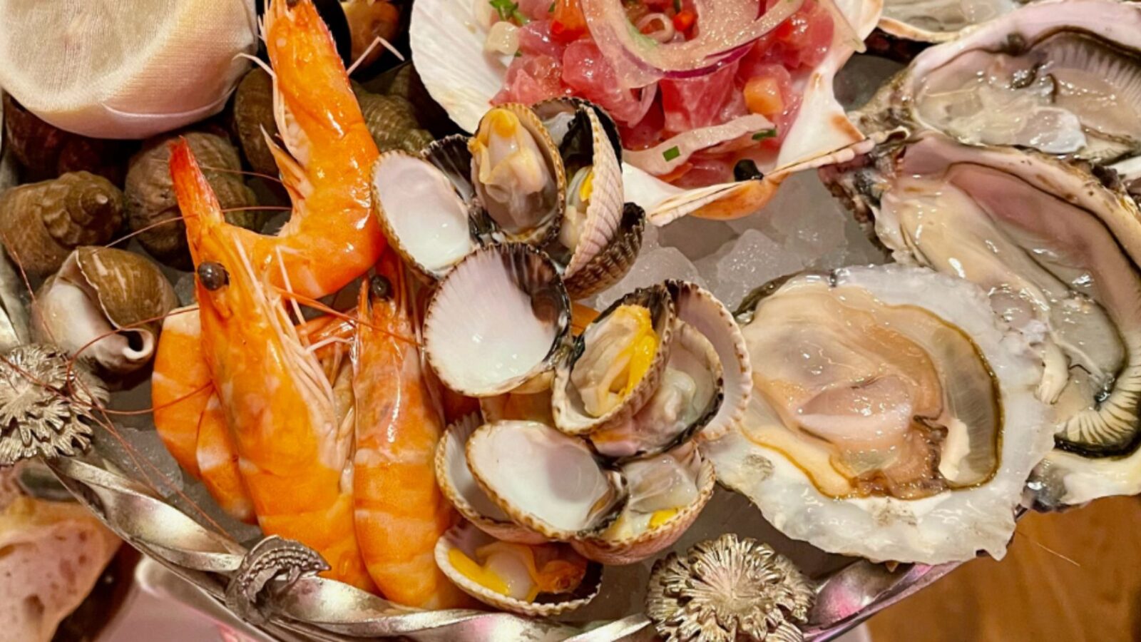 An ornate seafood tower made of a silver metal and topped with shrimp, escargot, and oysters.