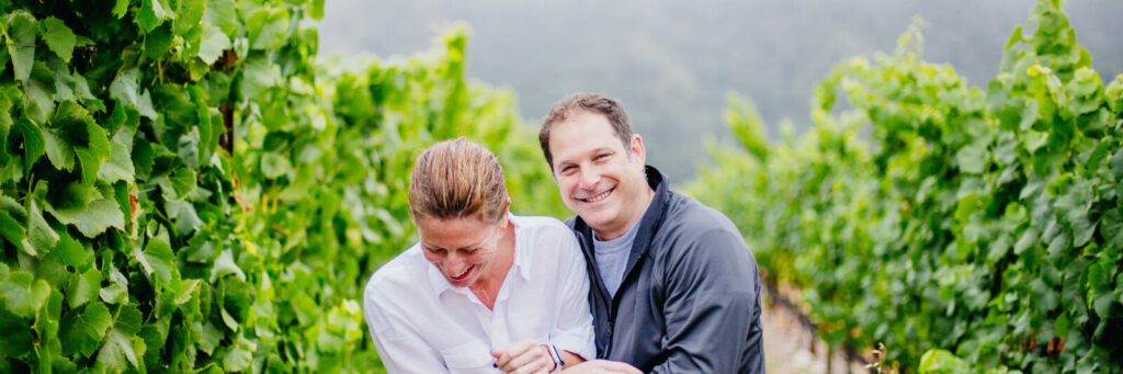 A couple holding each other and laughing in a vineyard of flourishing grape vines.