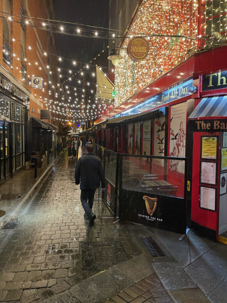 String lights hang over a cobblestone street with bars on either side of it.