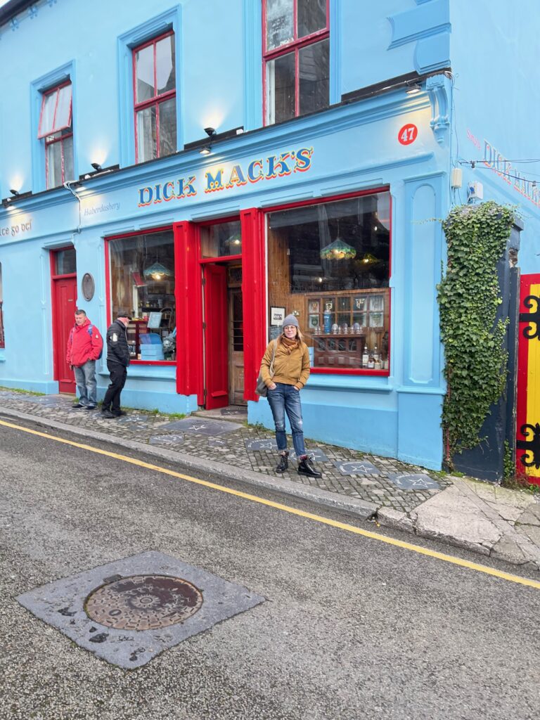 A woman smiles in front of a blue building with a sign that says "Dick Mack's" on it.