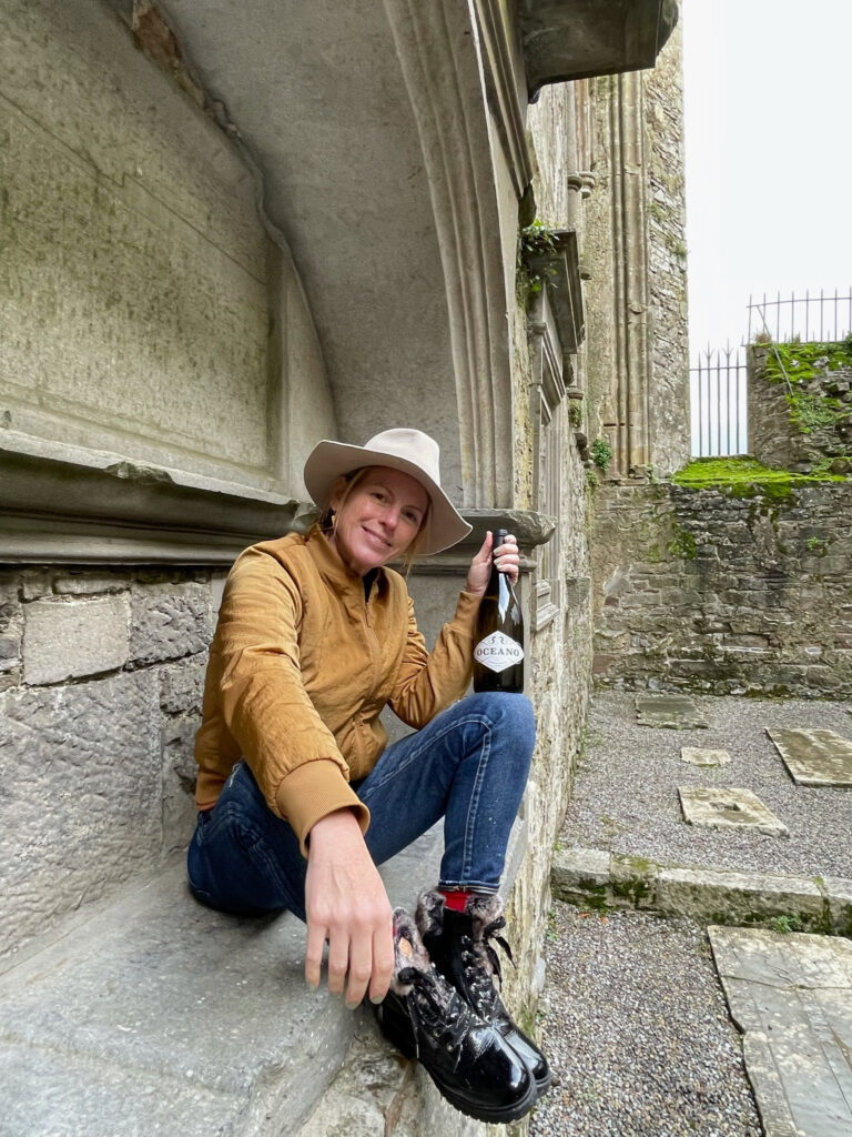 A woman in a yellow coat sits on a stone bench and smiles while holding a bottle of Oceano wine.