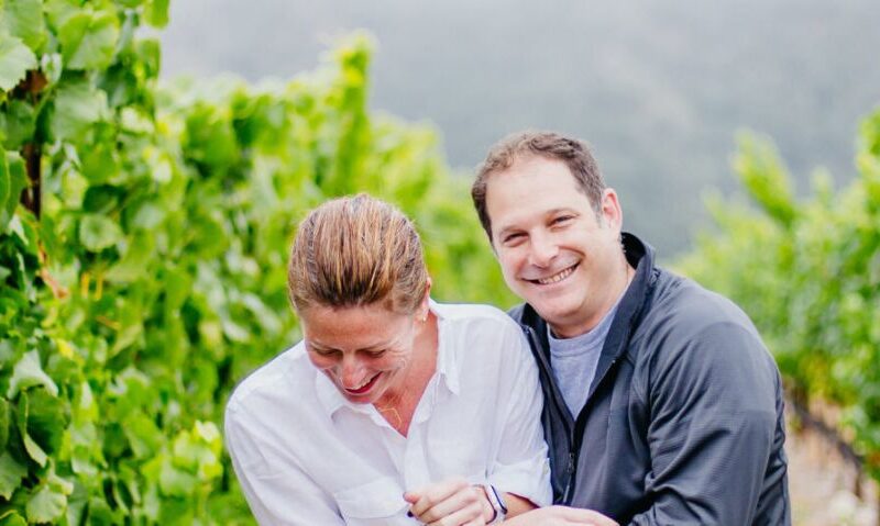 A couple holding each other and laughing in a vineyard of flourishing grape vines.