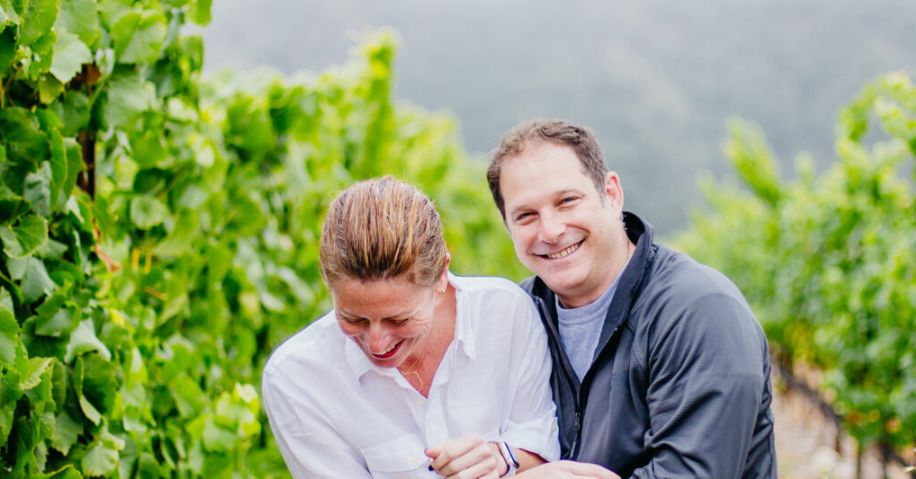 A couple holding each other and laughing in a vineyard of flourishing grape vines.