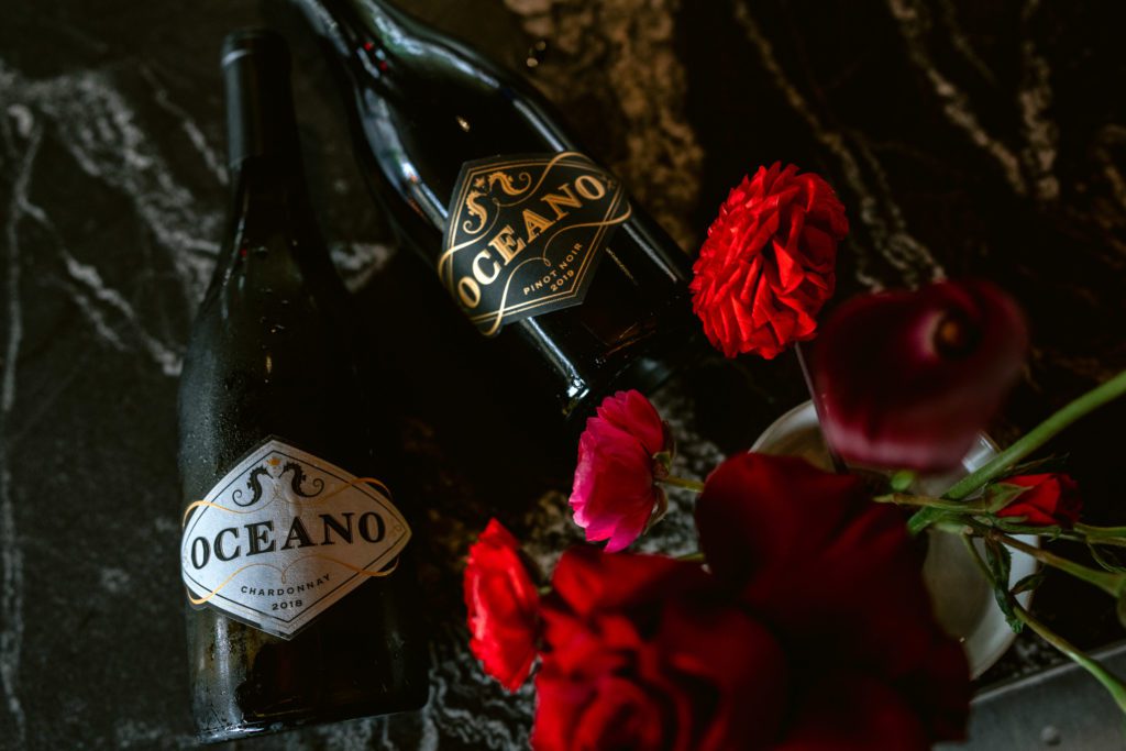 Two bottles of Oceano Wine: Chardonnay and Pinot noir, on a marble background with red flowers in the foreground.