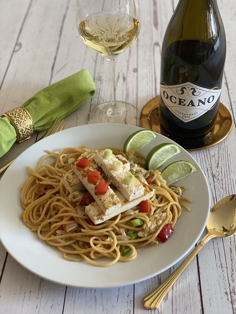 A plate with a tofu and noodle dish and three limes next to a wine bottle and green fabric napkin.