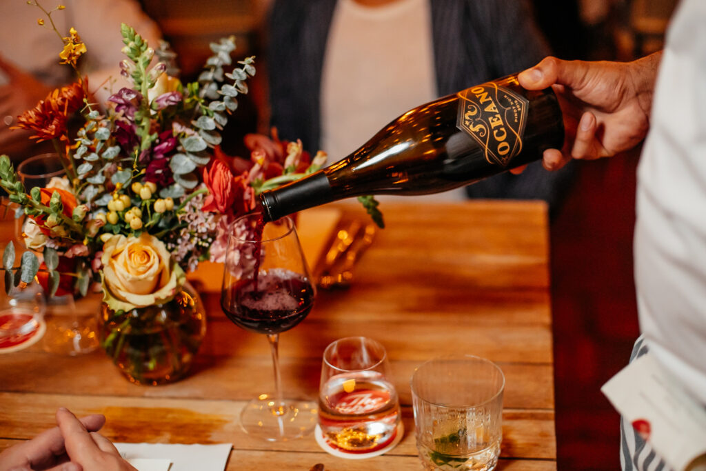 A man pouring a glass of red Oceano wine into a wine glass on a wooden table.