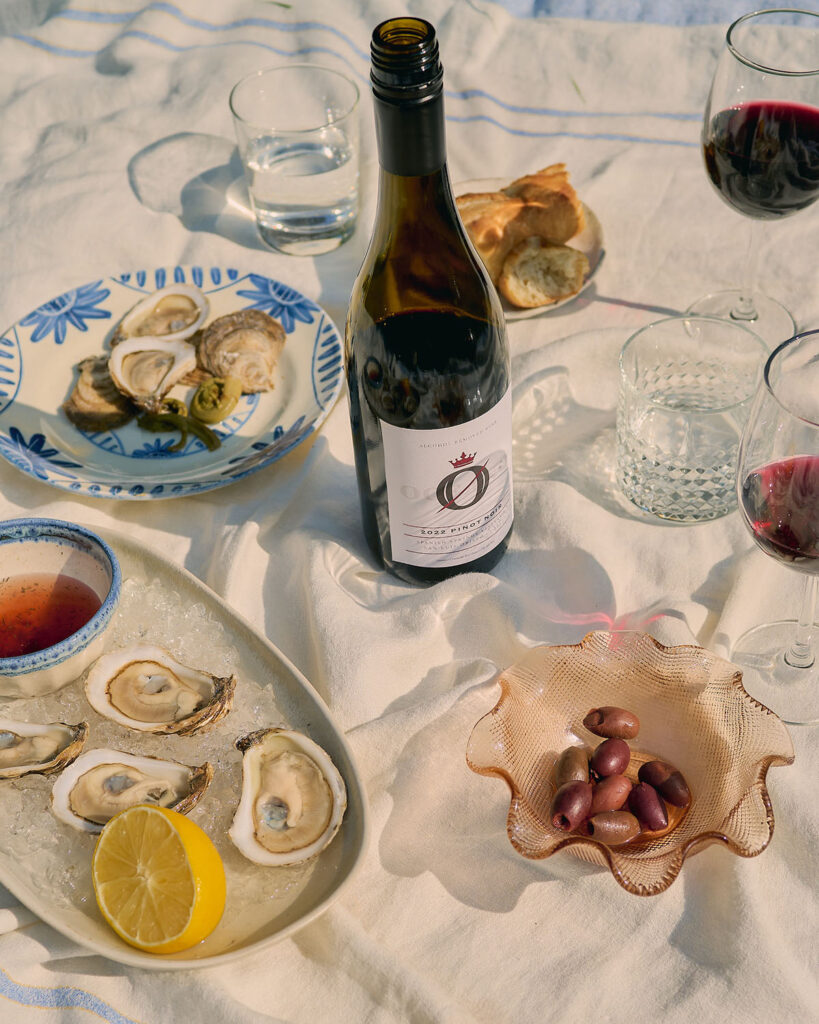 A picnic blanket with plates of mussels, olives, and bread and a bottle and glasses of alcohol free wine on it.