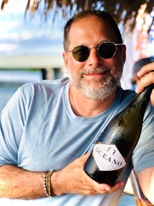 A man in a blue shirt and sunglasses smiles while holding an Oceano Wines wine bottle outside.