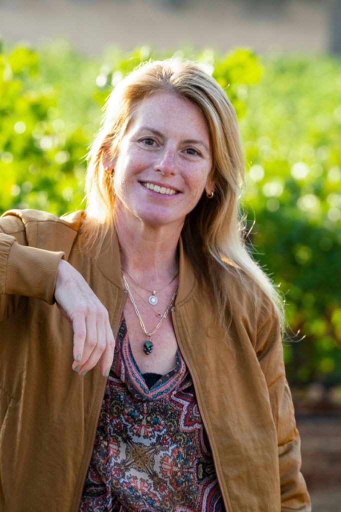 A woman in a patterned shirt and brown jacket smiles at the camera.